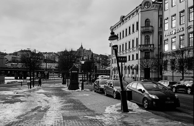 Parked cars and pedestrians beside ornate buildings on a sunlit city street