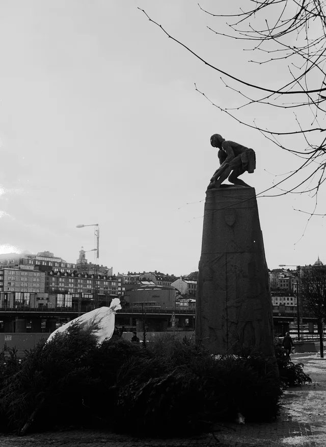 A crouching figure sculpture on a tall stone pedestal beside water