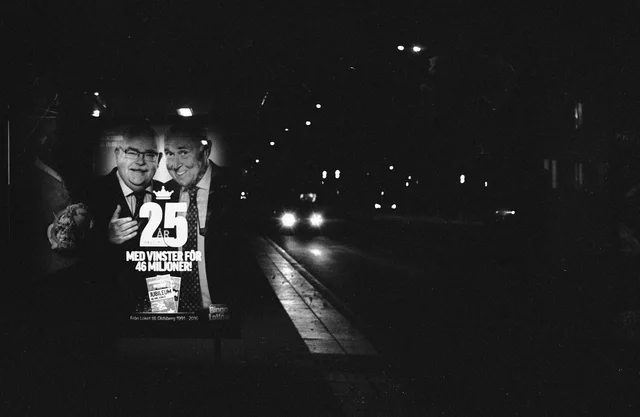 An illuminated advertising poster beside a dark road and distant headlights