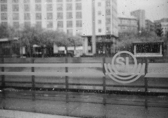 A rain-speckled window with a circular transit emblem and city buildings beyond