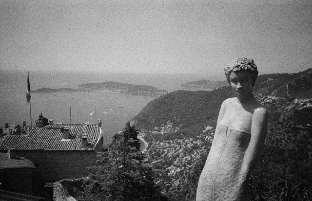 A weathered stone figure overlooking rooftops, hills, and a distant bay