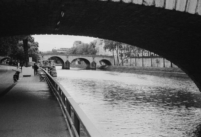 A riverside path passing beneath a bridge, with another arched bridge beyond