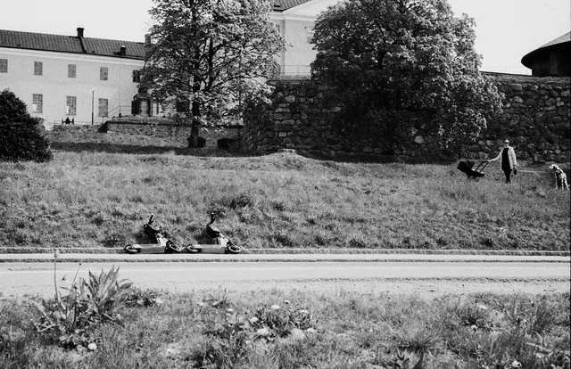 People resting and walking on a grassy riverbank below a stone wall and buildings