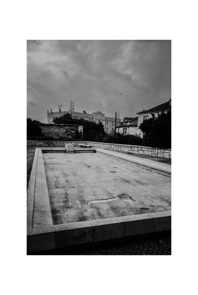 An empty rectangular stone basin in a paved square below distant buildings