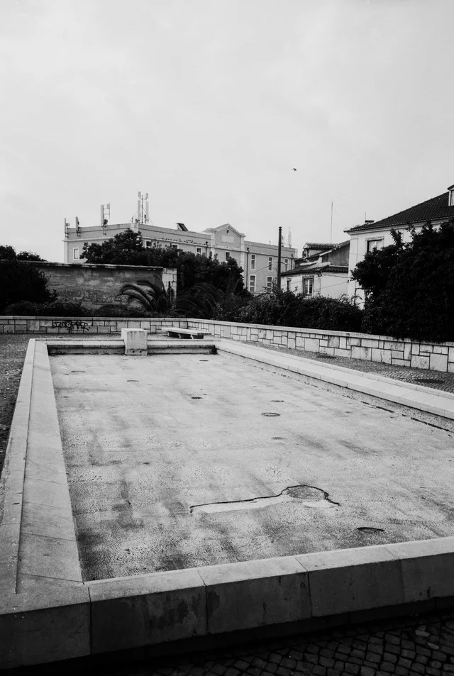 An empty rectangular stone basin in a paved square below distant buildings