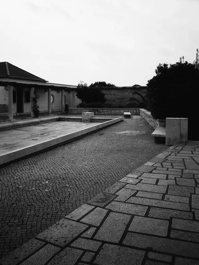 A long stone basin beside a paved walkway, a columned pavilion, and trees