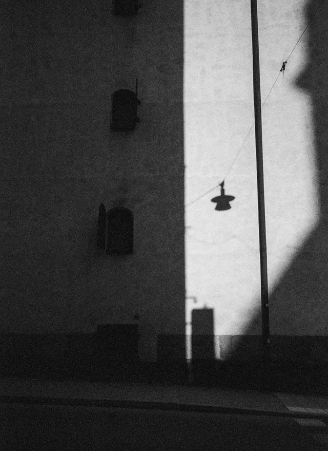 A hanging streetlamp silhouetted against a sunlit strip of a tall wall