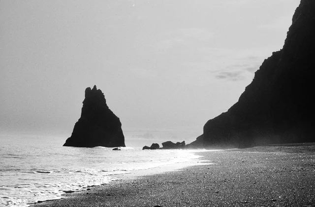 A pointed sea stack beside a dark beach and breaking waves