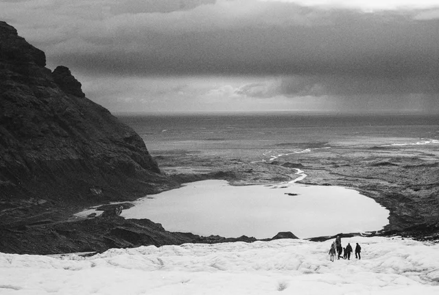 People crossing a snow-covered slope above a lake, with dark cliffs to the left