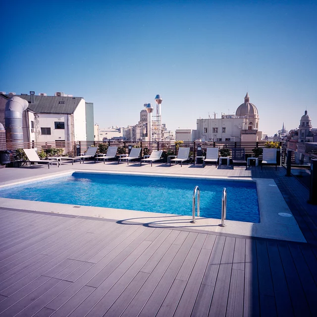 A bright blue rooftop swimming pool with loungers and a city skyline beyond